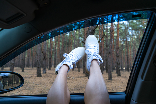 Legs of a young girl in white boots in a car window on a background of coniferous forest. Enjoyment, relaxation in the forest, lazy Sunday, tourism, travel, free time, rest, the concept of happiness.