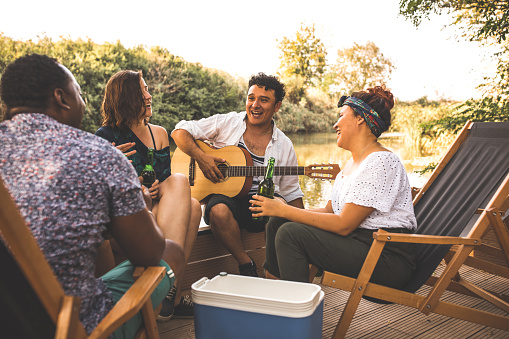 Group of happy young friends sitting by the river, singing, talking, playing guitar and drinking beer together.