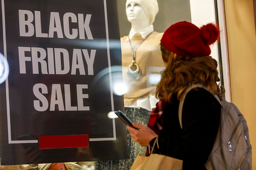 Selective focus shot of woman passing by a store window with a promotional poster that says ''Black Friday sale'' and using her smart phone.