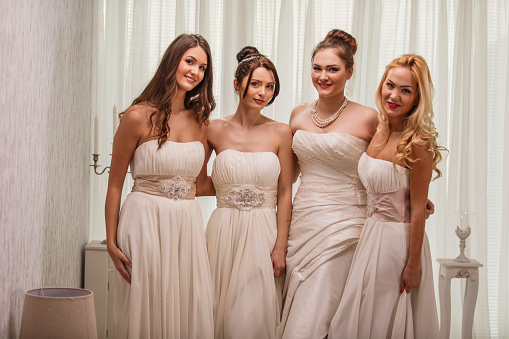 Three quarter length shot of four young brides in beautiful wedding dresses standing embraced and smiling at camera.