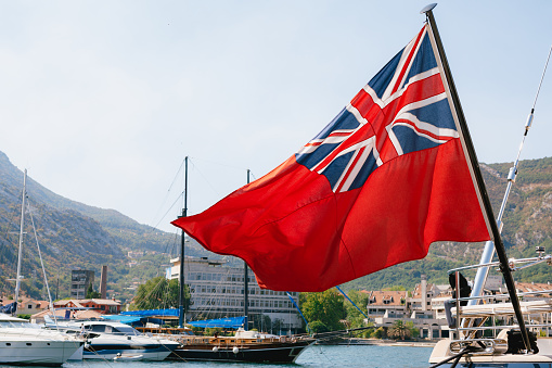 Close-up of the British Red Ensign aboard a ship with ships and mountains in the background. Red flag with the first version of Union Jack in the upper left corner. High quality photo
