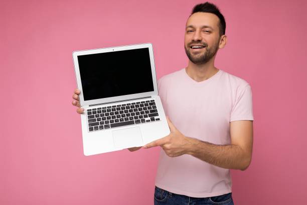 孤立したピンクの背景にtシャツでカメラを見てラップトップコンピュータを保持しているハンサムな笑顔のブルネットの男 - looking at camera smiling desk isolated ストックフォトと画像