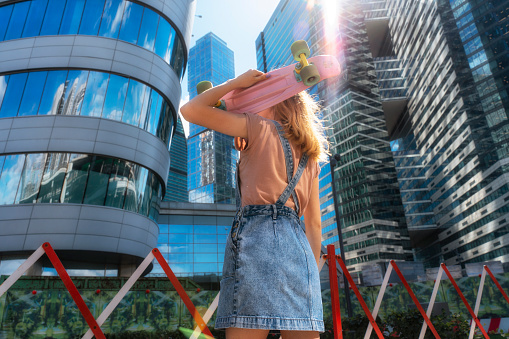 young girl 16 years old in jeans clothes with a skateboard looks at the quarter with skyscrapers in a big city on a sunny summer day with sunny highlights