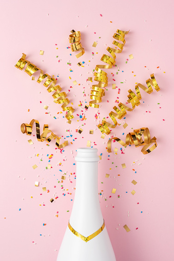 White champagne bottle with confetti and party streamers on pink background. Minimal party concept.