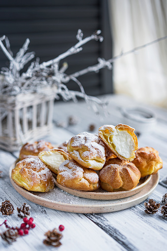 Homemade profiteroles stuffed with whipped white cream and powder sugar on wooden rustic table near window. Beautiful christmas or winter dessert. Copy space.