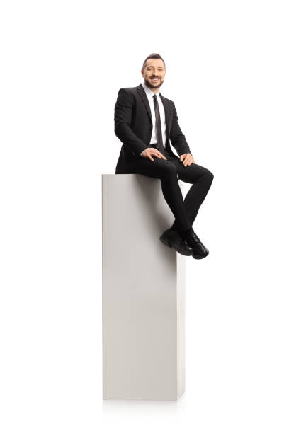 istock Man in a suit and tie sitting on a tall white column and smiling at camera 1295503142