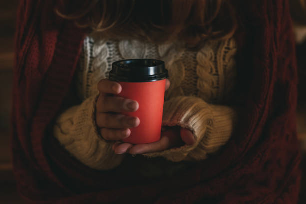 Woman holds in her hands red paper coffee cup. Woman wears white knitted woolen sweater and red plaid on the shoulders. Dark toned photography. Cozy theme. Selective focus.