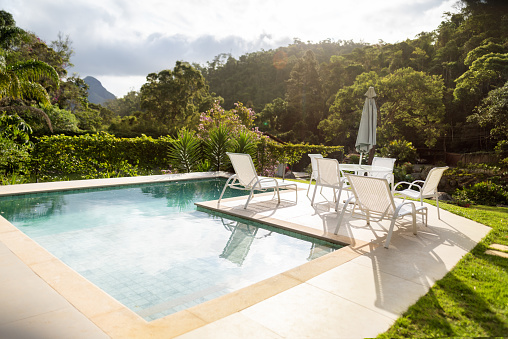 Comfortable loungers, chairs and a table sitting next to a swimming pool in the lush back yard on a sunny afternoon