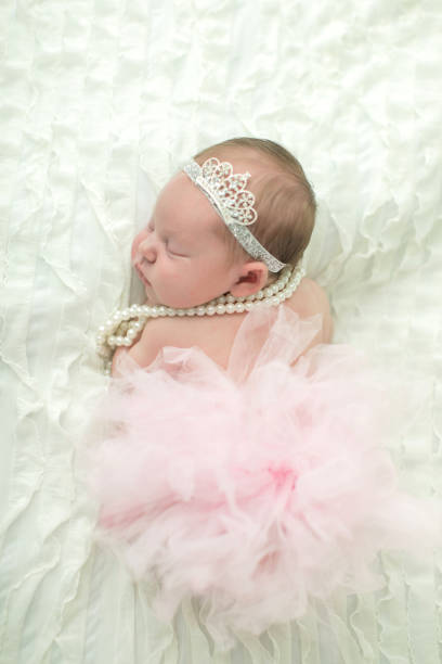 close up of a sweet newborn infant baby girl laying on a cream-colored neutral background with a crown on her head like a little princess royalty. - royal baby imagens e fotografias de stock