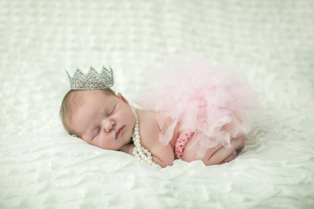 close up of a sweet newborn infant baby girl laying on a cream-colored neutral background with a crown on her head like a little princess royalty. - royal baby imagens e fotografias de stock