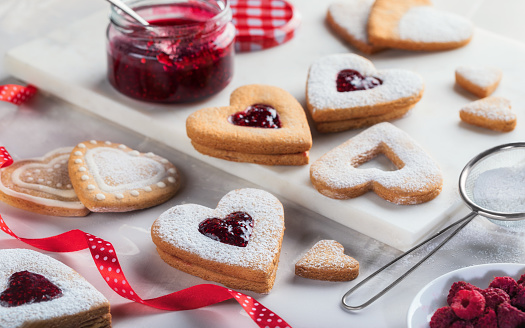 Heart shaped cookies with raspberry jam, sprinkled with powdered sugar on light gray background. Valentine's Day or Christmas biscuits making process.