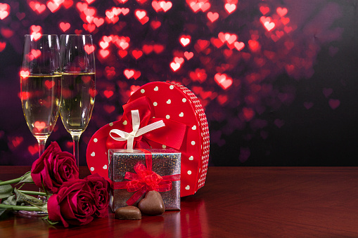 Valentine's Day. Composition: red heart-shaped gift box with bows, silver box with red bow, two champagne glasses, heart-shaped chocolates and three burgundy red roses.