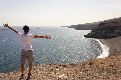 Young man with open arms on top of cliff enjoying ocean views in Lanzarote. Male tourist facing volcanic coast on sunny day in Canary Islands. Summer vacation, travel adventure concepts