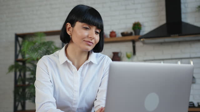 Lady in white blouse works on laptop at table in kitchen