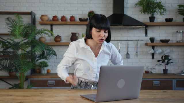 Emotional lady talks having videocall on laptop in kitchen