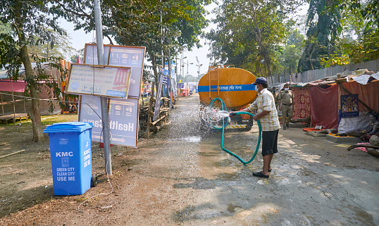 Babughat, Kolkata, 01/09/2021: A Kolkata corporation (KMC) vehicle water spraying dirt road, to keep the dust pollution under control inside the transit camp for Gangasagar Mela 2021. Several camps setup as resting place for pilgrims and sadhus are seen on both side of the road.