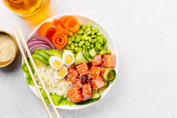 Photo of Poke bowl with salmon fish, rice, avokado, red onion, cucumber, edamame beans, carrot and quail eggs, sesame seedes. White background.