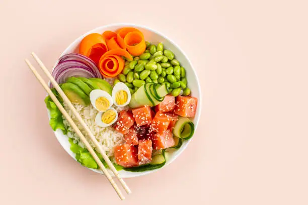 Photo of Poke bowl with salmon fish, rice, avokado, red onion, cucumber, edamame beans, carrot and quail eggs, sesame seeds. Light peach background.