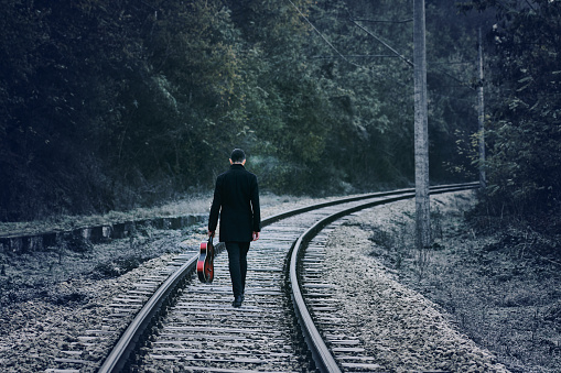 A young man stands on the rails with a guitar in his hands. Dressed in black