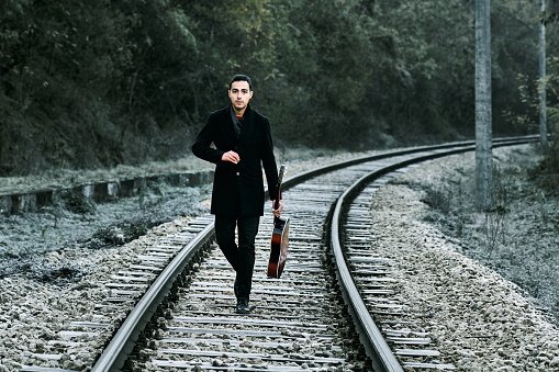 A young man stands on the rails with a guitar in his hands. Dressed in black