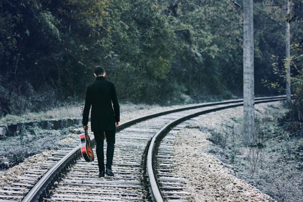 A young man stands on the rails with a guitar in his hands. Dressed in black