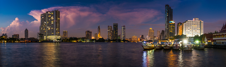 Panoramic view across the Chao Phraya river at sunset overlooked by the modern skyscrapers of Bangkok, Thailand’s vibrant capital city.