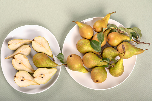 Group of pears on white plates and light green tablecloth. The pears are freshly brought from tree and have leaves. Two plates, one with whole pears and second with cut slices.