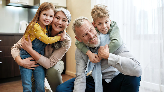 Happy family, grandparent with grandchildren spending time together, hugging and smiling at camera while standing in the kitchen at home. Younger and older generation