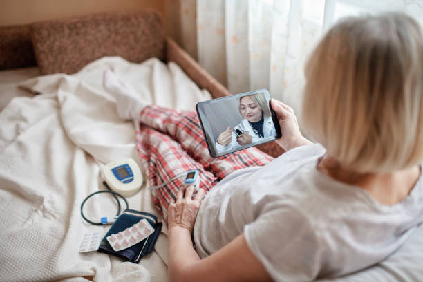 old woman in bed looking at screen of laptop and consulting with a doctor online at home, telehealth - controlo remoto imagens e fotografias de stock