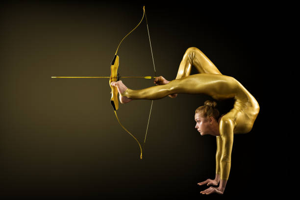 Archer Shooting by Legs with Gold Bow and Arrow. Flexible Gymnast aiming Target standing on Hand upside down. Goal Achievement Concept Archer Shooting by Legs with Gold Bow and Arrow. Flexible Gymnast aiming Target standing on Hand upside down. Goal Achievement Concept, Studio shot over Black background acrobatic activity stock pictures, royalty-free photos & images