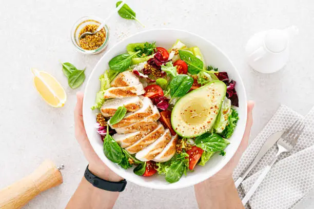 Photo of Grilled chicken meat and fresh vegetable salad of tomato, avocado, lettuce and spinach. Healthy and detox food concept. Ketogenic diet. Buddha bowl in hands on white background, top view