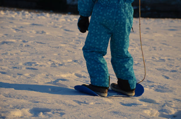 niño en invierno mono azul trata de montar una tabla de snowboard de plástico para niños. prueba la estabilidad y se aferra a la cadena. - mono ski fotografías e imágenes de stock