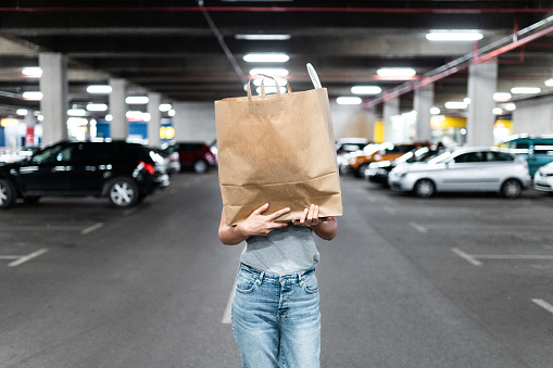 Woman in jeans and shirt walking across underground parking garage and carrying large paper bag in front of her