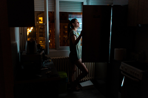 Young beautiful woman standing in kitchen and looking into fridge at night