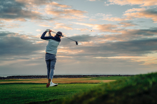 Male athlete teeing off at golf course against sky. Sportsman is practicing at sunset. He is swinging club.