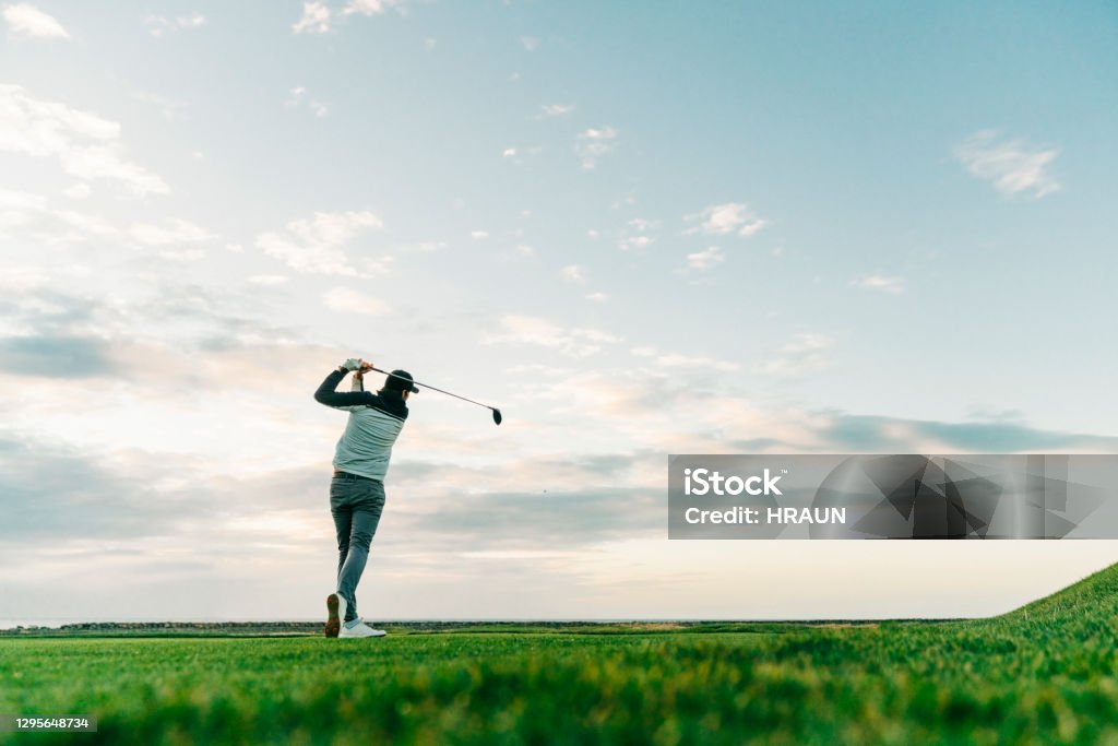 Male golfer swinging club at course during sunset Surface level view of sportsman taking a shot at course against sky. Rear view of male golfer is swinging club. He is practicing at sunset. Golf Stock Photo