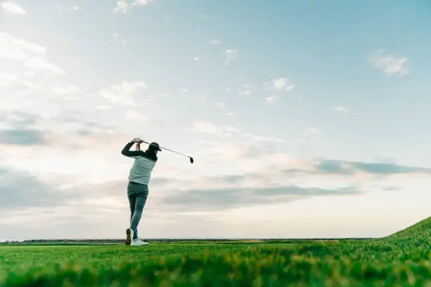 Surface level view of sportsman taking a shot at course against sky. Rear view of male golfer is swinging club. He is practicing at sunset.