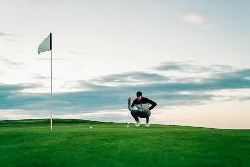 Full length of male golfer lining up ball and flag against sky. Sportsman is crouching while aiming on putting turf. He is playing at sunset.