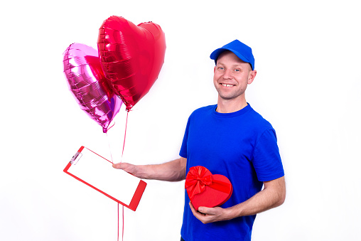 Courier in blue uniform holds red heart-shaped gift box and balloon for Valentine's Day. Home delivery of holiday gifts to your favorite women on Mother's and International Women's Day. I love you