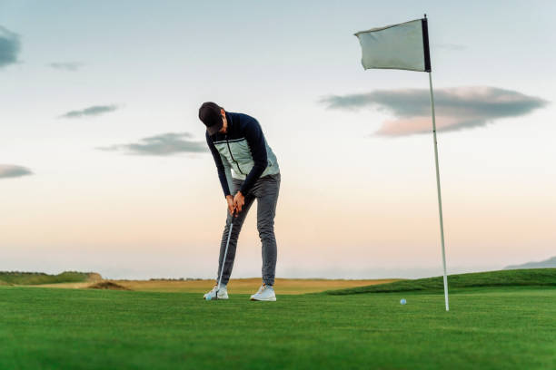 Sportsman taking a shot on golf putting turf Full length of sportsman taking a shot on putting turf against sky. Mid adult male golfer is at golf course. He is playing at sunset. putting golf stock pictures, royalty-free photos & images