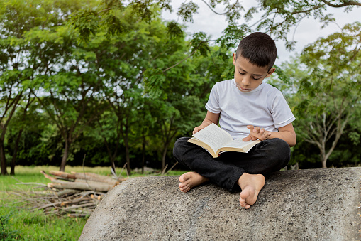 Latino boy 7-12 years old reading a book sitting on a concrete pipe on a sidewalk barefoot, sunny day with blue sky, the concept of education and rela