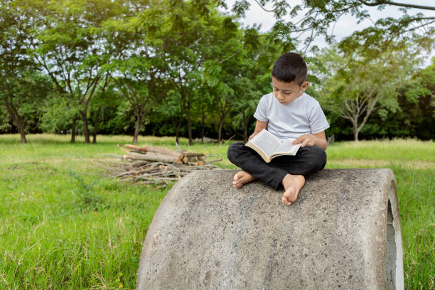 istock Boy reading a book sitting on a concrete pipe on a sidewalk 1295650619