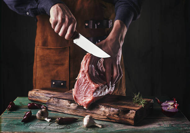 um cara de avental de couro está cortando carne crua. o açougueiro corta as costelas de porco - meat steak raw market - fotografias e filmes do acervo