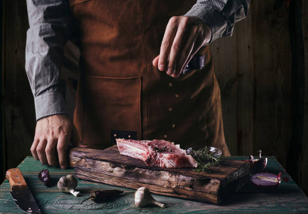a guy in a leather apron sprinkles black pepper on a steak with a bone. - sirloin steak fotos imagens e fotografias de stock