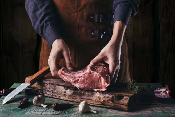 um cara de avental de couro está cortando carne crua. o açougueiro corta as costelas de porco - rib eye steak beef cutting board meat - fotografias e filmes do acervo