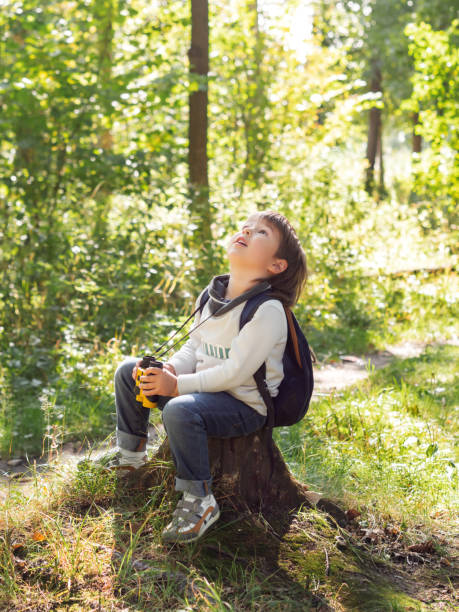 istock Little explorer on hike in forest. Boy with binoculars sits on stump. Outdoor leisure activity for children. Summer journey for young tourist. 1295652956