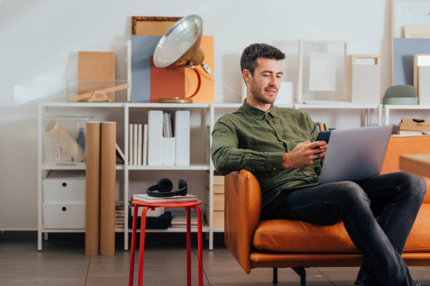 istock New Normal Concept: Young Businessman Working from Home on his Smartphone and his Laptop PC 1295653979