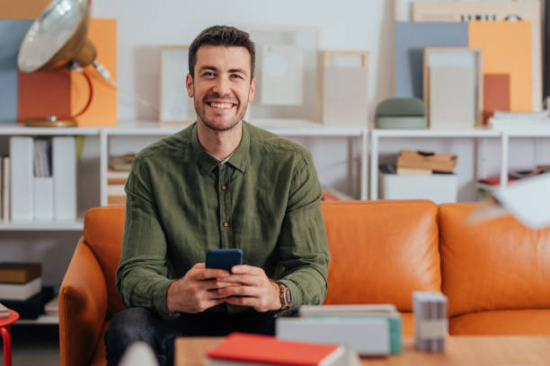 retrato de un empresario moderno sonriente sentado en la oficina y uso de su teléfono inteligente para comunicarse en línea - white green indoors studio shot fotografías e imágenes de stock