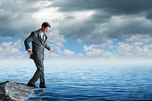 A man dressed in a suit gingery dips the toe of his shoe into the water as he stands on a rock. The dark clouds above begin to clear as blue skies appear on the horizon.