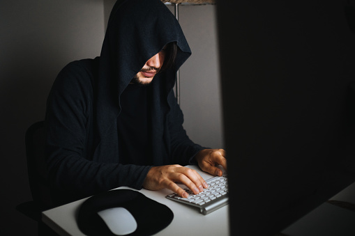 Photo of adult man to hide his identity and using computer for hacking.The background is dark and atmosphere is futuristic. He is using a keyboard